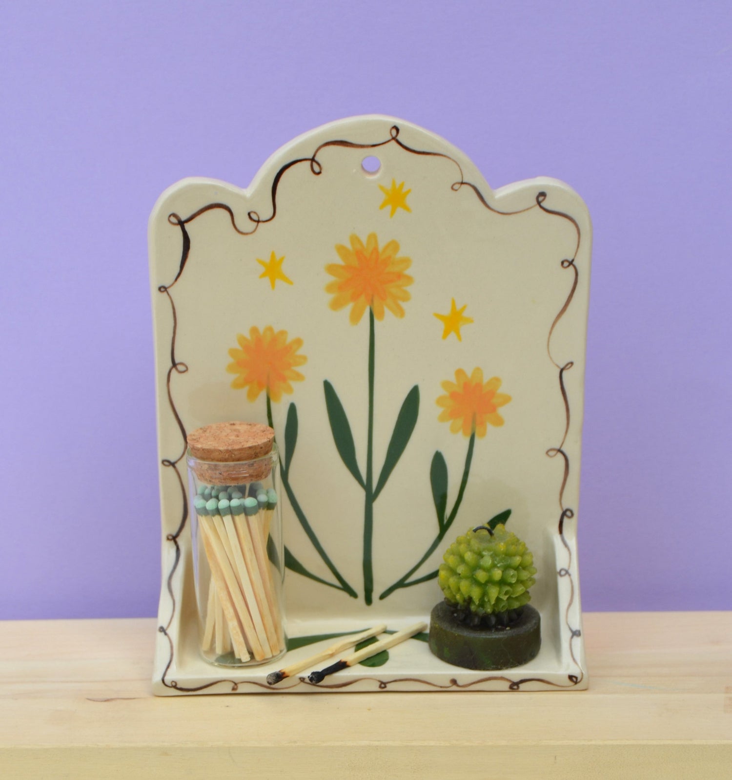 Ceramic hanging altar with illustrative dandelion design. Shelf has a jar of matches and a little cactus displayed on it.