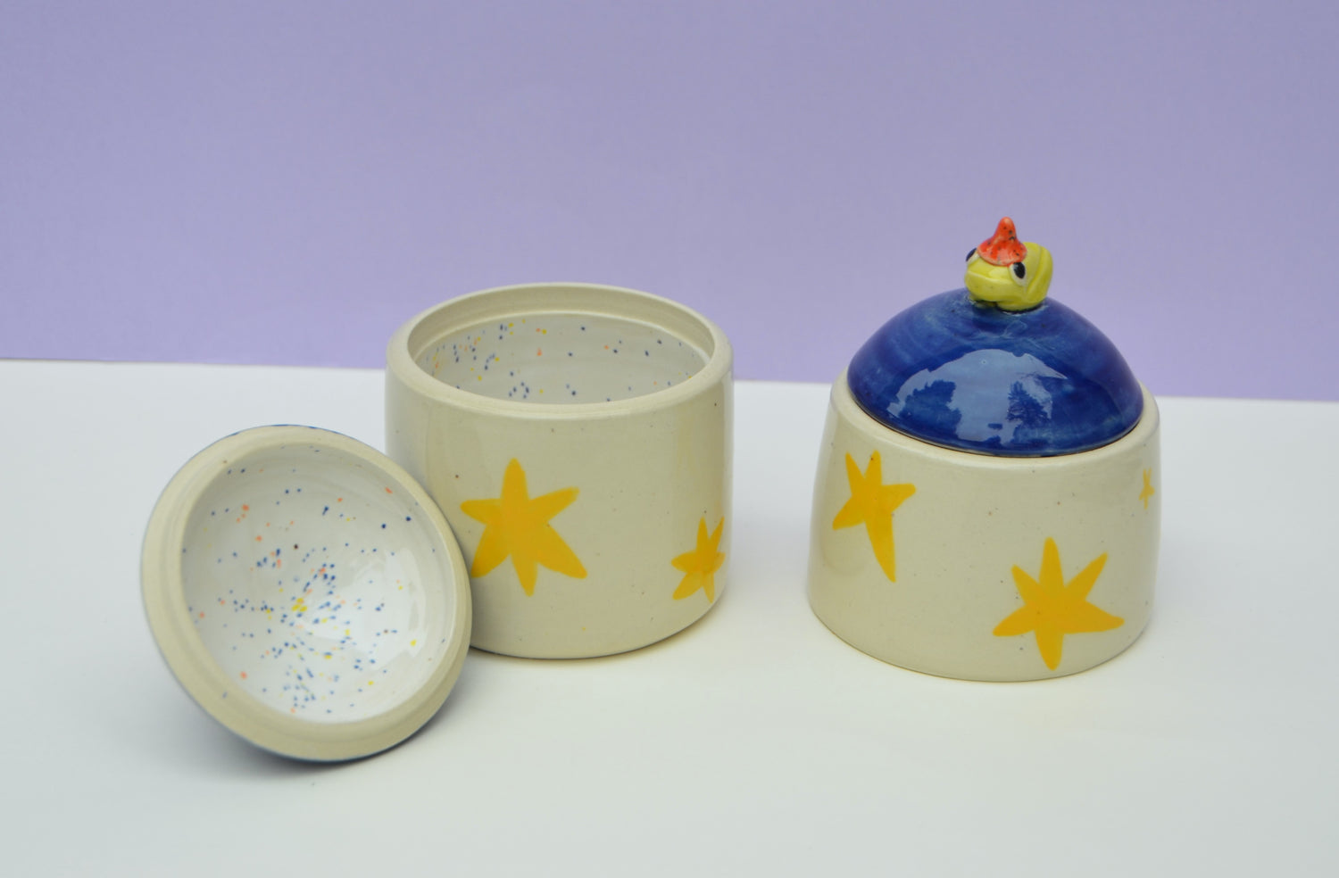 two ceramic jars, jar on the left is open showing off its speckled white interior. The jar on the right is lidded with a dark blue lid that features a small ceramic frog wearing a wizard hat. both jars have bright yellow stars decorating the outside