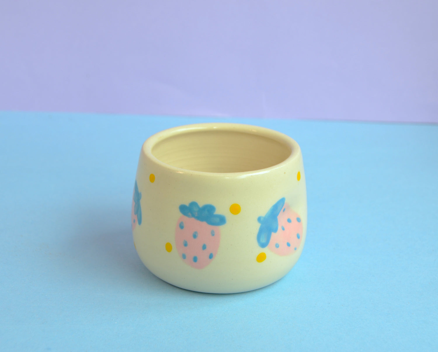 ceramic cup with yellow dots and light pink strawberries with light blue leaves. Cup features an indented side for a comfortable grip