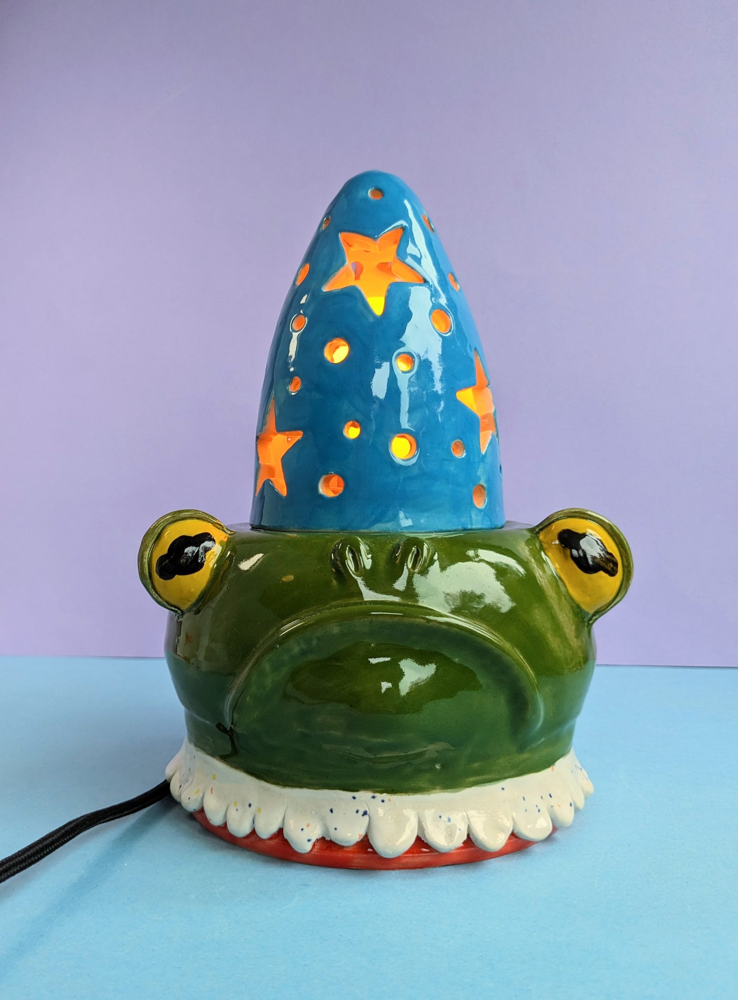 Ceramic Frog wizard lamp featuring a light blue hat with glowing stars, a dark green frowning frog base that has a white ruffle collar.